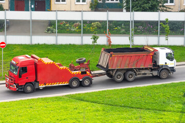 Powerful big red semi evacuation truck tractor tows with attached broken evacuated truck with body driving on city highway. - obrazy, fototapety, plakaty