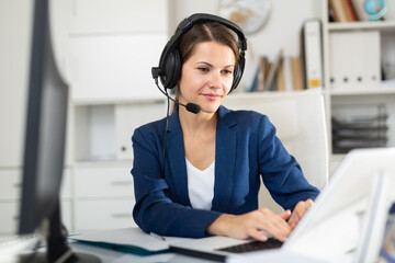 Portrait of positive woman call center operator receiving calls and using laptop in office
