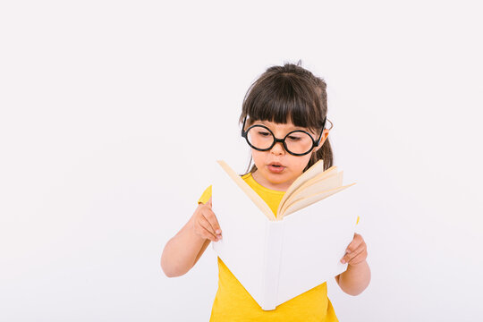 Surprised little girl, wearing yellow t-shirt and round black glasses holding an open book in her hands and reading, on white background