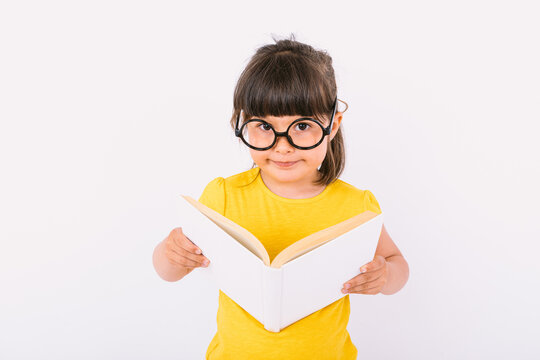 Little girl smiling, wearing yellow t-shirt and round black glasses, holding an open book in her hands and looking at camera, on white background