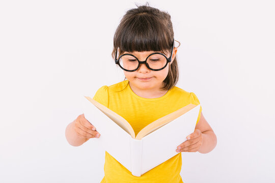 Smiling little girl, wearing yellow t-shirt and round black glasses holding an open book in her hands and reading, on white background