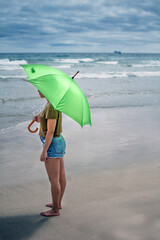 Joven muchacha de pie frente al mar mientras sostiene un paraguas verde en una playa de fina arena