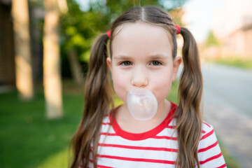 Cute child girl with two ponytails looking at camera and inflating bubble with chewing gum outdoor. Kids blowing bubble gum