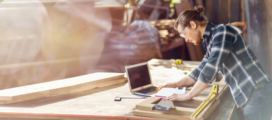 young hipster carpenter man working with computer laptop in workshop . craftsman wearing safety glasses checking order of clients or learning online - obrazy, fototapety, plakaty