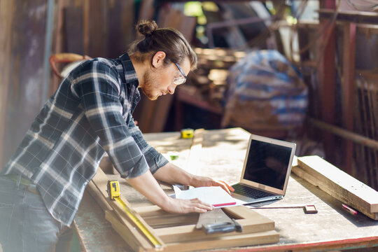 young hipster carpenter man working with computer laptop in workshop . craftsman wearing safety glasses checking order of clients or learning online