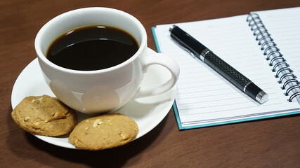Black coffee in a white cup with cookies on wooden table, And notebook with pen beside, Coffee break time and writing idea concept, Close up