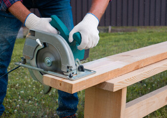 A man builder with an electric hand-held circular saw saws a wooden board on the lawn. DIY in the backyard.