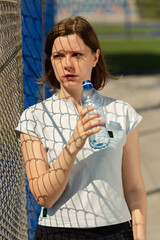 Woman drinking water after fitness training on the outdoor sports ground. Healthy lifestyle with...