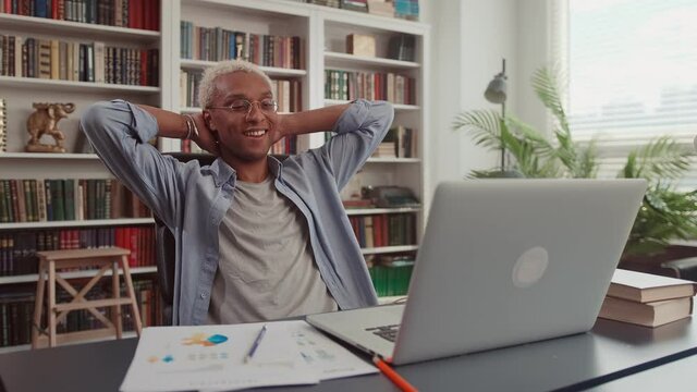 Calm millennial man in glasses sit relax at home office workplace take nap or daydream. Happy relaxed African American young male rest in chair distracted from computer work, relieve negative emotions