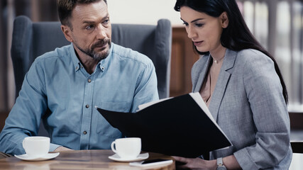 businessman looking at woman with paper folder near cups of coffee on table