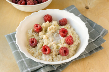 Milk barley porridge with butter and raspberries in white bowl on wooden background