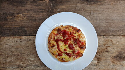 Top view of small homemade pizza with ham Mozzarella cheese, cheese and tomato sauce Sprinkle on a thick pizza crust. Served in a round white plate on a wooden table. Is a dish that often cook at hom
