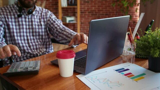 Businessman in wheelchair works on laptop using calculator on table with tables, closeup. Concept of using digital gadgets by people with disabilities