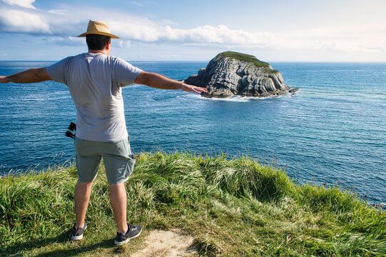 Hombre con sombrero de paja y brazos en cruz contemplando la isla del Castro en la costa quebrada de Cantabria, España