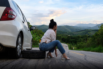 Young Asea woman sitting near the car for calling for help on the public road in the forest area at mountain and sky background