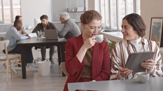 Waist-up of Caucasian woman wearing red formal suit, drinking hot tea and chatting with her female Mixed-Race friend using tablet computer, sitting at desk in office