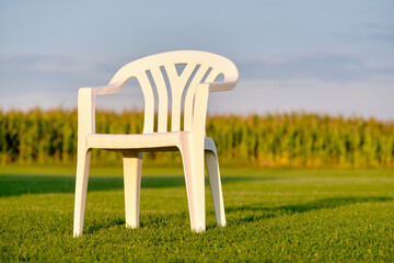 Empy white plastic garden chair standing on a green lawn in front of a cornfield in evening light....