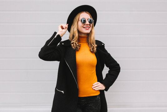 Fashionable portrait of stylish happy smiling young woman having fun female model posing wearing a black coat, round hat on city street on gray background