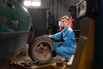 Fototapeta na wymiar A woman auto mechanic sits near a disassembled car, dressed in overalls and safety glasses, holds a wheel and looks into the camera.