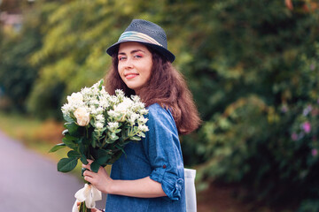 Portrait of a young beautiful woman in a straw hat holding a bouquet of white roses. Plants and trees on the background. The concept of March 8 and Mother's Day