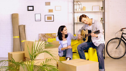 cheerful man playing acoustic guitar near happy woman on couch in new home