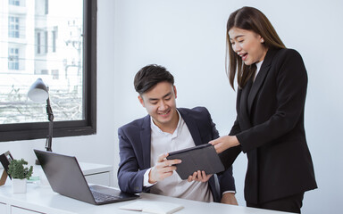 Asian young handsome business man and beautiful secretary wearing formal suits and smiling to each others while a woman presenting her project to the boss by using tablet in the office.