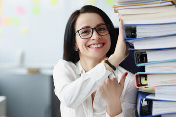 Young woman with glasses holding many folders with documents in office