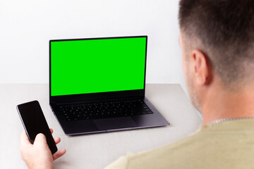 a man with a phone in his hand is sitting in front of a laptop with a green chromakey mockup on the monitor, the concept of office work, remote work, marketing, training, coaching