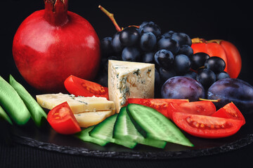 A slice of blue cheese on a black flat slate plate with fruits: pomegranate, plums, grapes,...