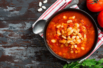 White bean soup with vegetables in a black bowl on a dark wooden background. Greek bean soup with...