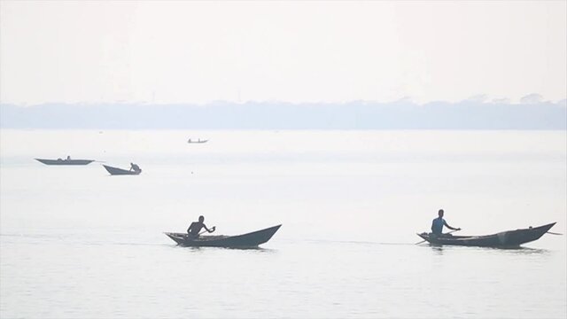 Men sitting boats paddle along the Padma river in hazy sunshine.
