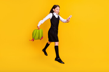 Full length body size photo schoolgirl jumping with rucksack using smartphone isolated vibrant yellow color background
