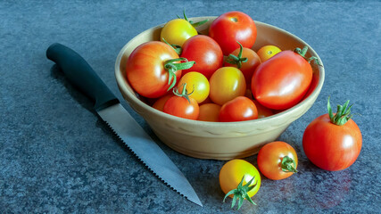 Freshly harvested organic mixed tomatoes. In a simple brown glazed bowl, sitting on blue grey marble. With others on surface and a serrated edged kitchen knife. Interior landscape with selective focus - 450535433