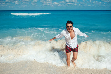 Attractive playful tourist man in white shirt and sunglasses enjoying day on the beach and running thru sea waves