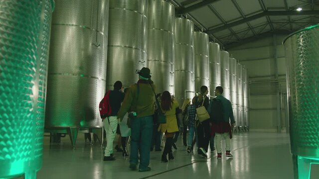 Group of tourists visited winemaker or alcohol production factory . Female guide showing big steel barrels . Fermentation of wine , brandy or whiskey . People in winery . Huge conveyors . Slow motion