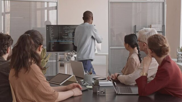 Medium long of young African IT specialist making presentation, showing computer code on screen to board of directors sitting at conference table