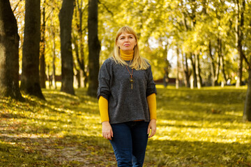 Defocus pretty blond 40s woman standing in yellow autumn park. Happy beautiful lady. Women wearing grey pullover, yellow turtleneck, pendant, jeans. Fall park with leaves. Out of focus