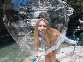 A girl looks through a huge soap bubble at another bursting bubble on the background of rocks and a river