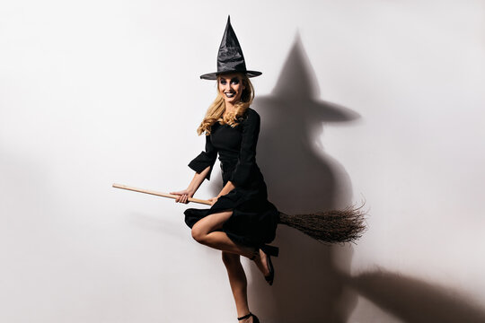 Ecstatic young woman in witch hat having fun in carnival. Indoor photo of elegant caucasian girl sitting on magic broom.