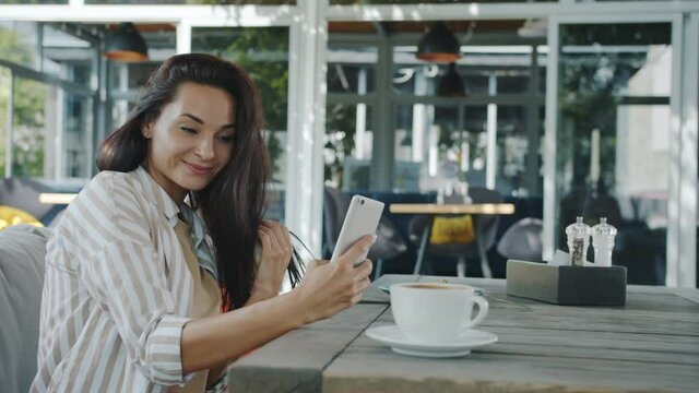 Cheerful young lady is taking selfie with smartphone camera posing in outdoor cafe alone having fun with modern device. Photograph and lifestyle concept.