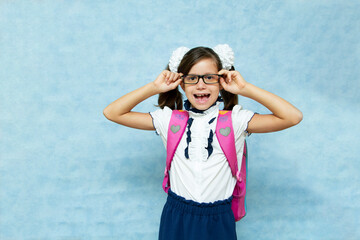 Funny dark-haired girl in glasses in school clothes with a backpack on her back on a blue background. Preparation for school. Place for your text.
