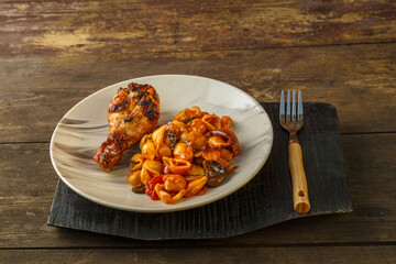 Seashell pasta in a tomato with a chicken leg baked on a grill on a wooden stand on a wooden table next to a fork.