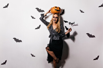 Graceful woman in black skirt dancing at halloween party. Smiling blonde girl holding pumpkin on white background.