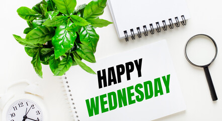 On a light background, there is a white alarm clock, a magnifying glass, a green plant and a notebook with the words HAPPY WEDNESDAY.