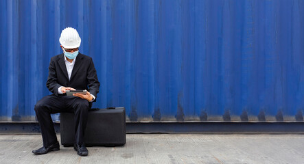 asian manager businessman wearing face mask to protect form covid or coronavirus sitting on bag and working seriously on tablet with worrying business crisis situation from covid at construction site.