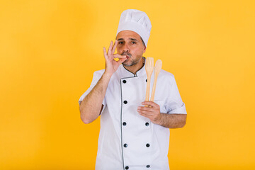 Chef cook wearing a white kitchen cap and jacket holds a spoon and a wooden fork and makes the gesture of delicious food with the other hand, on yellow background