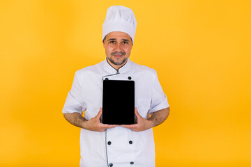 Chef cook wearing white kitchen jacket and hat, holding a tablet and pointing a finger, on yellow background