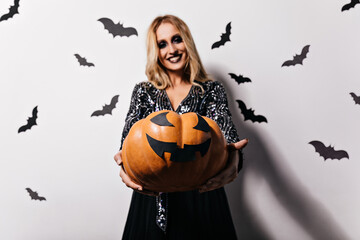 Excited fair-haired woman with black makeup holding halloween pumpkin. Studio shot of smiling...