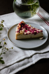 A slice of cake with farmer cheese and raspberries with some field flowers on a small plate on a piece of table cloth and wooden table
