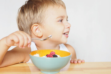 a blond boy with brown eyes in a white T-shirt eats cottage cheese with berries at a wooden table..
